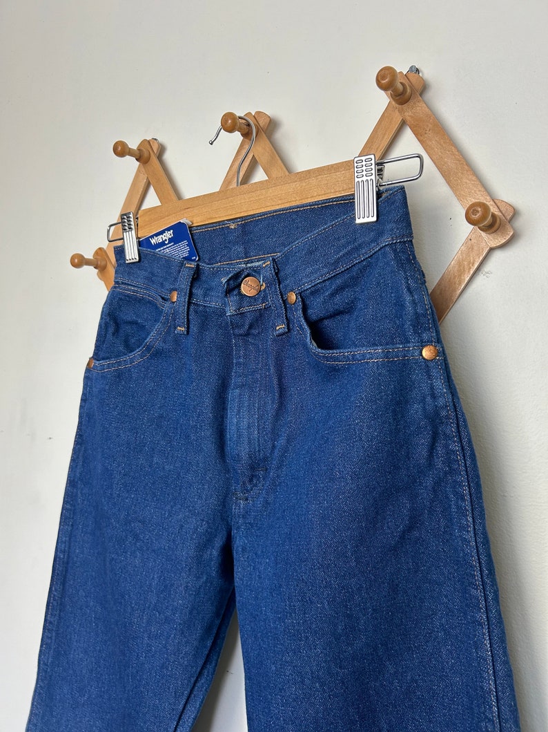 Puede incluir: Vaqueros Wrangler azul oscuro colgados en un perchero de madera. Los vaqueros tienen un dise&ntilde;o cl&aacute;sico de cinco bolsillos con remaches color cobre y cierre de bot&oacute;n. Una etiqueta azul con el logotipo de Wrangler est&aacute; adjunta.