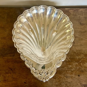 May include: A silver-colored, shell-shaped serving dish with a scalloped edge. The dish has a ribbed design and ornate floral detailing along the bottom edge. The reflective surface of the dish creates a bright, elegant appearance.