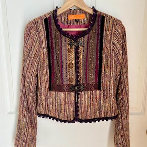 Puede incluir: Chaqueta de tweed multicolor con un panel frontal decorativo. La chaqueta presenta una mezcla de tonos morados, dorados y marrones, con detalles de terciopelo negro y cierres ornamentados. La chaqueta tiene cuello redondo y mangas largas.