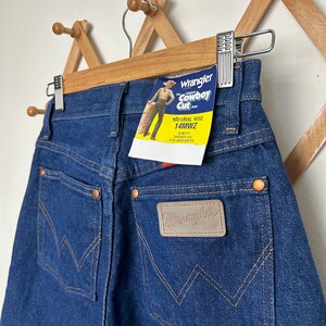 Puede incluir: Vaqueros Wrangler Cowboy Cut azul oscuro con tiro natural y corte entallado, pernera c&oacute;nica. Los vaqueros tienen un parche de cuero con el logo de Wrangler y una etiqueta que dice "Wrangler Cowboy Cut". Los vaqueros est&aacute;n colgados en una percha de madera.