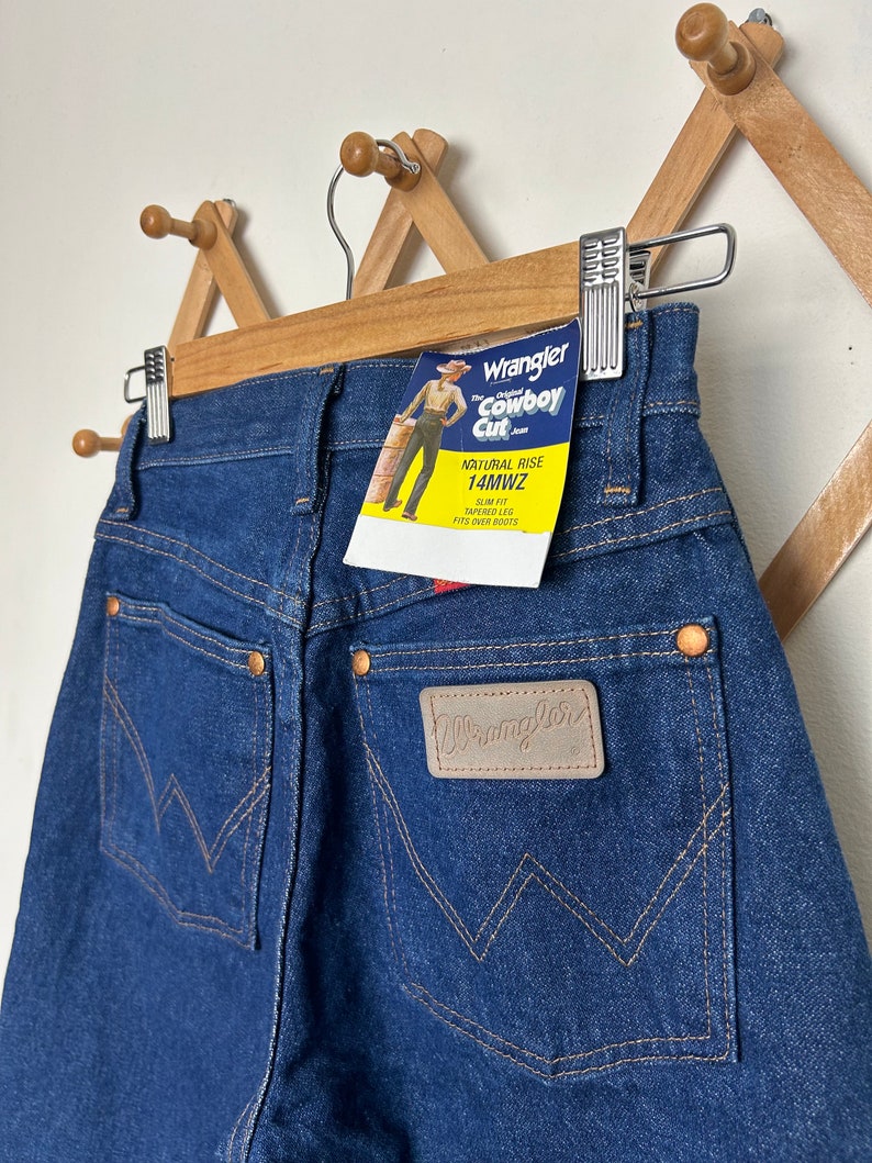 Puede incluir: Vaqueros Wrangler Cowboy Cut azul oscuro con tiro natural y corte entallado, pernera c&oacute;nica. Los vaqueros tienen un parche de cuero con el logo de Wrangler y una etiqueta que dice "Wrangler Cowboy Cut". Los vaqueros est&aacute;n colgados en una percha de madera.