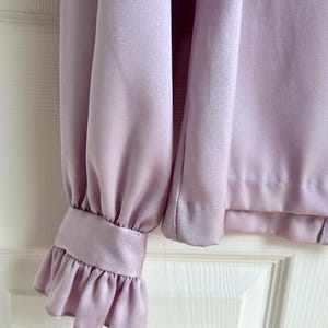 May include: A close-up of a lilac-colored blouse sleeve. The sleeve features a ruffled cuff and a smooth, silky texture. The blouse is made of a lightweight fabric, and the design is simple and elegant.