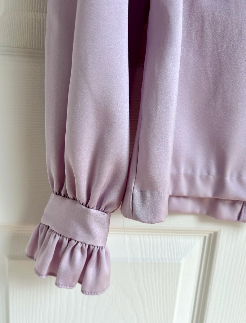 May include: A close-up of a lilac-colored blouse sleeve. The sleeve features a ruffled cuff and a smooth, silky texture. The blouse is made of a lightweight fabric, and the design is simple and elegant.