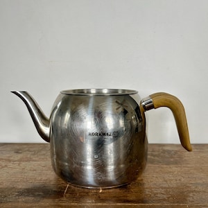 May include: A stainless steel teapot with a curved spout and a wooden handle. The teapot has a round body and the brand name "KORKMAZ" is visible on the side. The teapot is sitting on a wooden surface.