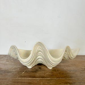 May include: A large, cream-colored ceramic clam shell-shaped bowl. The bowl has a wavy, textured design and sits on a wooden surface. The bowl is a decorative piece, suitable for coastal or nautical decor.