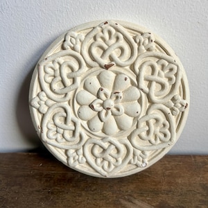 May include: A white ceramic wall decor with a circular design featuring a central flower surrounded by intricate knotwork patterns.