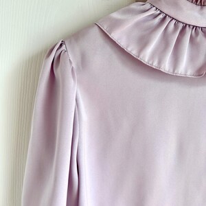 May include: A light purple blouse with a high ruffled collar and long sleeves. The fabric has a silky sheen, and the shoulder has a slight puff detail. The blouse is a solid color and appears to be made of a lightweight material.