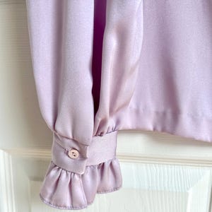 May include: Close-up of a lilac-colored blouse sleeve. The sleeve features a buttoned cuff with a small, round button and a ruffled edge. The fabric has a silky sheen.