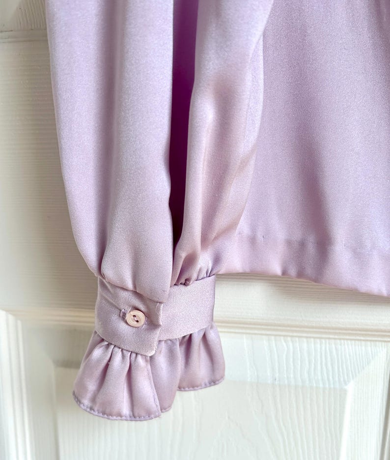 May include: Close-up of a lilac-colored blouse sleeve. The sleeve features a buttoned cuff with a small, round button and a ruffled edge. The fabric has a silky sheen.