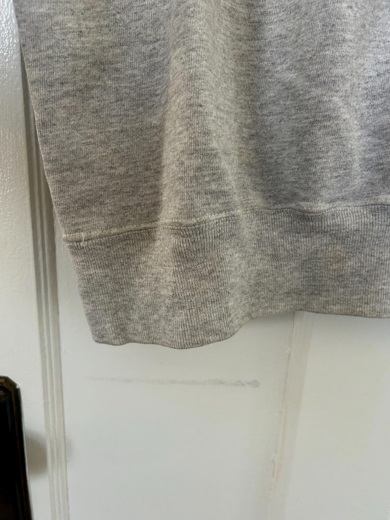 Pode incluir: Close-up da bainha de um moletom cinza. O tecido &eacute; um tecido de malha macia e texturizada. A bainha &eacute; canelada e costurada com uma costura vis&iacute;vel. O moletom &eacute; de cor s&oacute;lida e neutra.