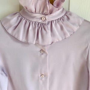 May include: A light purple blouse with a ruffled collar and button-down front. The blouse has long sleeves and a soft, silky texture. The collar is the focal point, with multiple layers of delicate ruffles.