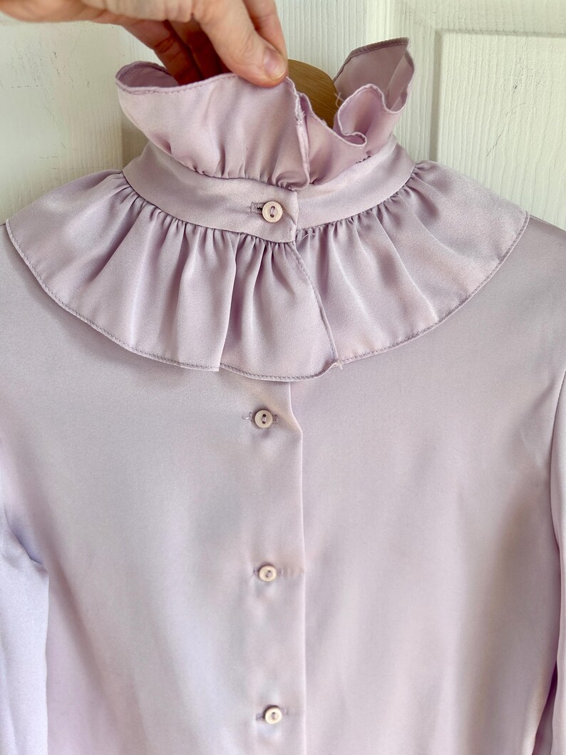 May include: A light purple blouse with a ruffled collar and button-down front. The blouse has long sleeves and a soft, silky texture. The collar is the focal point, with multiple layers of delicate ruffles.