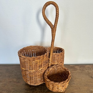 Pode incluir: Um conjunto de três cestos de vime tecido em castanho claro, com uma pega. Os cestos são de tamanhos diferentes e têm um aspeto natural e rústico. A pega é arqueada e os cestos são feitos de vime.