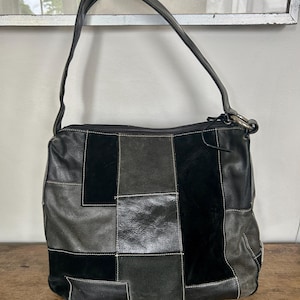 Puede incluir: Un bolso de hombro confeccionado con un mosaico de paneles de cuero y ante en negro y gris oscuro. El bolso cuenta con una sola correa, un cierre de cremallera superior con tirador de metal y costuras blancas visibles que delinean las secciones.