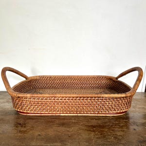 May include: A rectangular woven rattan tray featuring a natural light brown finish, a flat base, and two curved handles on opposite sides. The tray is constructed with a tight, uniform weave pattern throughout the sides and the bottom surface.