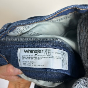 Puede incluir: Primer plano de unos vaqueros Wrangler de mezclilla azul. Los vaqueros tienen una etiqueta blanca con el logotipo de Wrangler y las instrucciones de lavado. Los vaqueros son 100% algod&oacute;n.