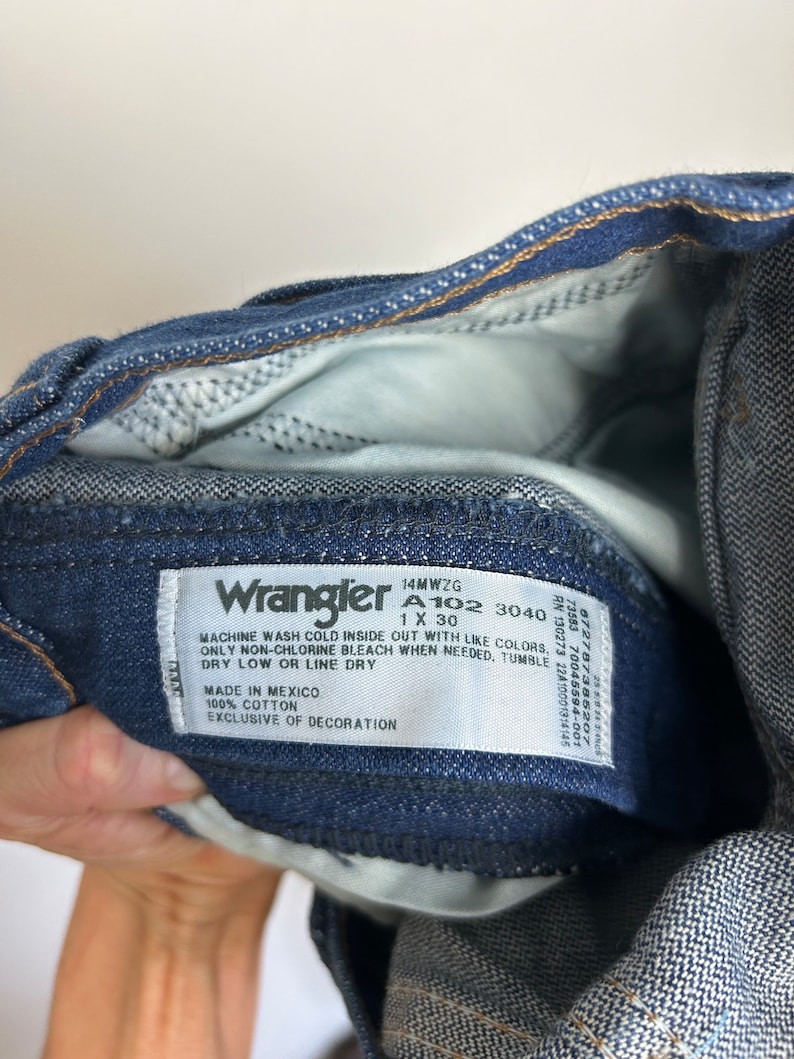 Puede incluir: Primer plano de unos vaqueros Wrangler de mezclilla azul. Los vaqueros tienen una etiqueta blanca con el logotipo de Wrangler y las instrucciones de lavado. Los vaqueros son 100% algod&oacute;n.
