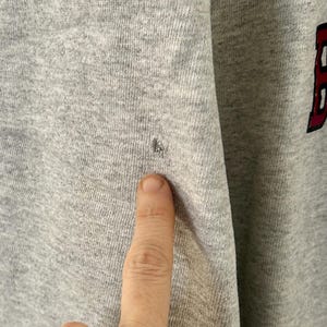Pode incluir: Close-up de um moletom cinza com uma pequena mancha escura. O tecido &eacute; texturizado e parece ser uma mistura de algod&atilde;o. A letra "B" em vermelho com uma contorno preto &eacute; vis&iacute;vel no lado direito da imagem.