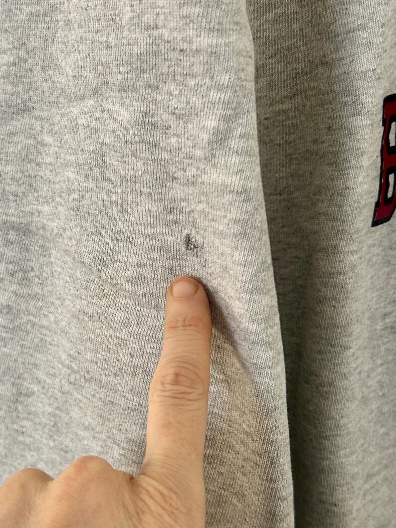 Pode incluir: Close-up de um moletom cinza com uma pequena mancha escura. O tecido &eacute; texturizado e parece ser uma mistura de algod&atilde;o. A letra "B" em vermelho com uma contorno preto &eacute; vis&iacute;vel no lado direito da imagem.