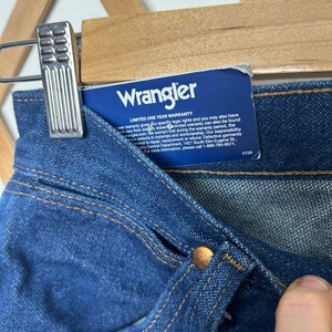 Puede incluir: Primer plano de unos vaqueros de mezclilla azules con un bot&oacute;n de color cobre y detalles de costura. Una etiqueta azul con el logotipo de Wrangler y la informaci&oacute;n de la garant&iacute;a est&aacute; adjunta. Los vaqueros cuelgan de una percha de madera.