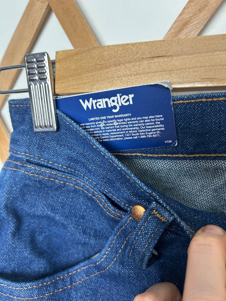 Puede incluir: Primer plano de unos vaqueros de mezclilla azules con un bot&oacute;n de color cobre y detalles de costura. Una etiqueta azul con el logotipo de Wrangler y la informaci&oacute;n de la garant&iacute;a est&aacute; adjunta. Los vaqueros cuelgan de una percha de madera.