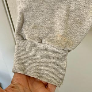 Pode incluir: Close-up de uma manga de moletom cinza com punho canelado. O tecido mostra sinais de desgaste e descolora&ccedil;&atilde;o, com manchas vis&iacute;veis perto do punho. A manga est&aacute; ligeiramente enrugada.