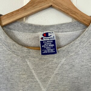 Pode incluir: Um moletom Champion X-Large cinza com gola redonda. A etiqueta diz "Champion Authentic Athletic Apparel" e "Assembled in Mexico". O moletom est&aacute; pendurado em um cabide de madeira.
