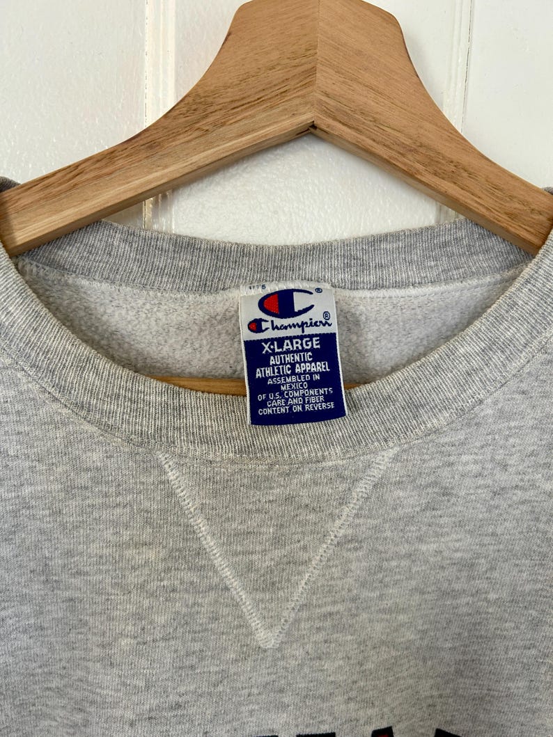 Pode incluir: Um moletom Champion X-Large cinza com gola redonda. A etiqueta diz "Champion Authentic Athletic Apparel" e "Assembled in Mexico". O moletom est&aacute; pendurado em um cabide de madeira.