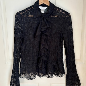 以下が含まれることがあります： 黒いレースのブラウス。長袖とフリル付きのフロントが特徴です。襟付きで、木製のハンガーにかかっています。「Allison Taylor」のブランド名がラベルに表示されています。