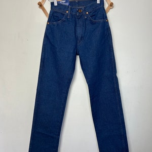 Puede incluir: Vaqueros de mezclilla azul oscuro con pernera recta. Los vaqueros tienen un dise&ntilde;o cl&aacute;sico de cinco bolsillos, remaches de cobre y cierre de bot&oacute;n y cremallera. Una etiqueta azul y blanca con la marca "Wrangler" est&aacute; adjunta.