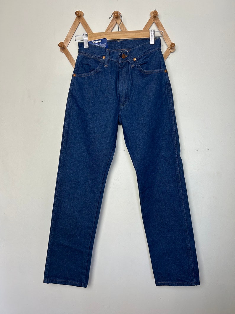 Puede incluir: Vaqueros de mezclilla azul oscuro con pernera recta. Los vaqueros tienen un dise&ntilde;o cl&aacute;sico de cinco bolsillos, remaches de cobre y cierre de bot&oacute;n y cremallera. Una etiqueta azul y blanca con la marca "Wrangler" est&aacute; adjunta.