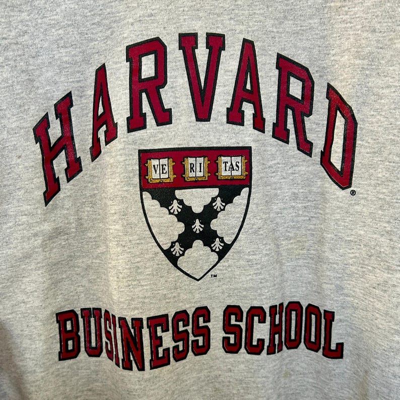 Pode incluir: Close-up de um moletom cinza com as palavras "HARVARD" em letras vermelhas em bloco arqueadas na parte superior. Abaixo, as palavras "BUSINESS SCHOOL" est&atilde;o em letras vermelhas em bloco. Um escudo com as palavras "VE RI TAS" est&aacute; no centro.