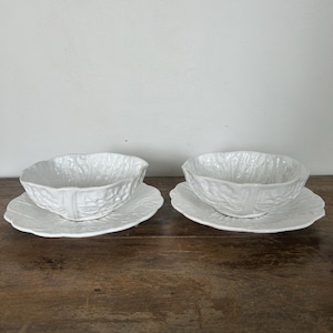 May include: Two white ceramic bowls and saucers. The bowls are shaped like cabbage leaves and sit on matching leaf-shaped saucers. The set is displayed on a dark wooden surface, creating a contrast with the bright white tableware.