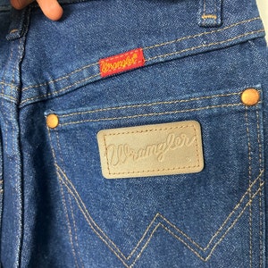 Puede incluir: Primer plano de vaqueros de mezclilla azules con el logotipo de Wrangler en un parche rojo y una etiqueta de cuero. Los vaqueros tienen remaches de color cobre y un dise&ntilde;o cl&aacute;sico de cinco bolsillos. La costura es de color beige contrastante.