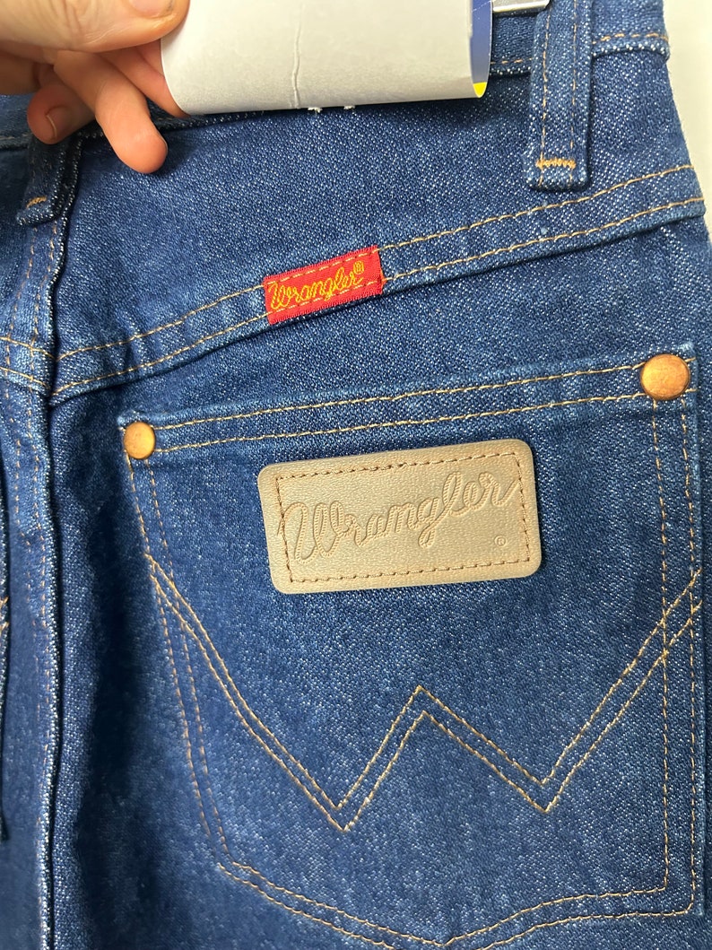 Puede incluir: Primer plano de vaqueros de mezclilla azules con el logotipo de Wrangler en un parche rojo y una etiqueta de cuero. Los vaqueros tienen remaches de color cobre y un dise&ntilde;o cl&aacute;sico de cinco bolsillos. La costura es de color beige contrastante.