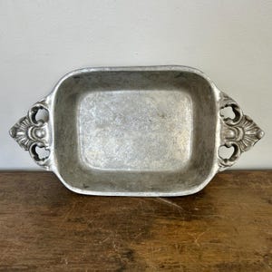 Puede incluir: Una bandeja rectangular de metal plateado con asas decorativas ornamentadas. La bandeja tiene un aspecto ligeramente desgastado, lo que sugiere que podría ser vintage o antigua. Las asas presentan un diseño floral estilizado.