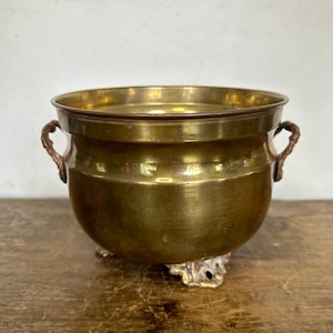 Op de afbeelding: Een gepolijste koperen pot met een rond lichaam, een brede rand en twee kleine decoratieve handvatten aan de zijkanten. De pot wordt ondersteund door drie sierlijke, getextureerde voetjes die de basis verhogen, met een warme gouden afwerking.