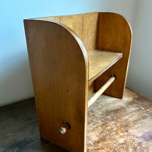 May include: A wooden wall-mounted paper towel holder with a shelf. The holder is made of light brown wood and has a rounded top. The shelf is also made of wood and is located above the paper towel holder. The holder has a wooden knob for dispensing the paper towels.