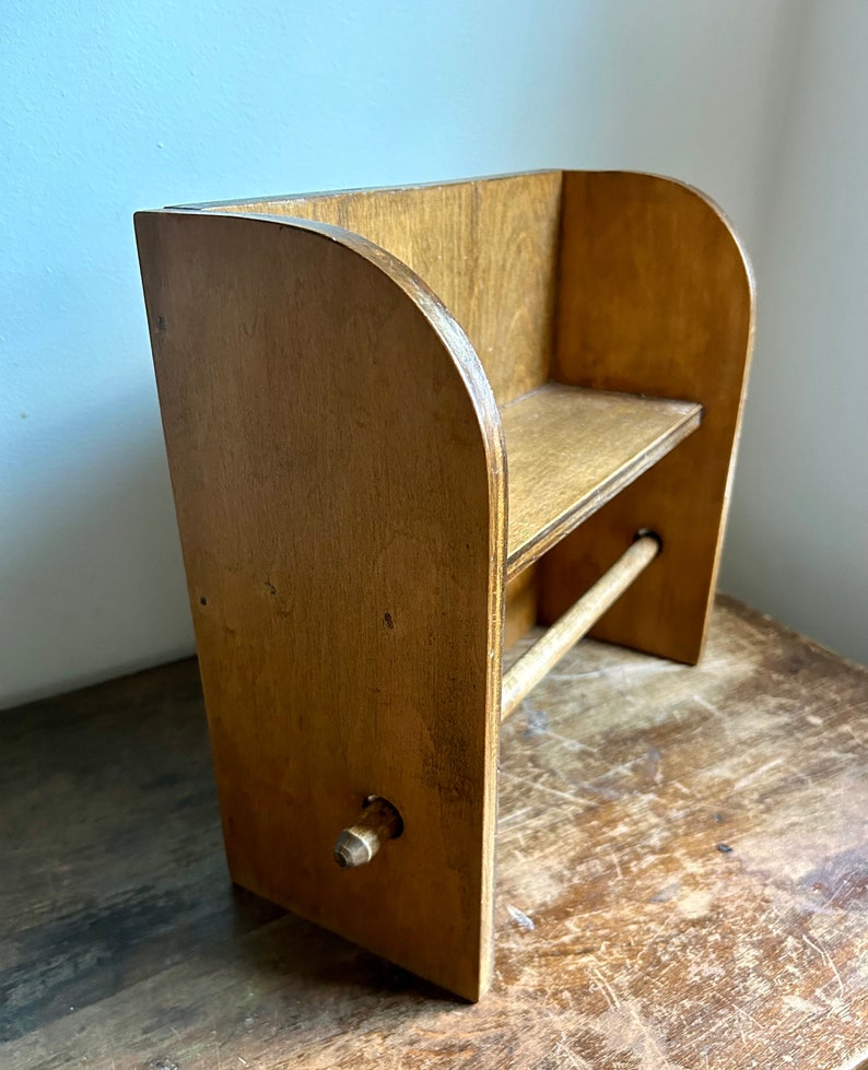 May include: A wooden wall-mounted paper towel holder with a shelf. The holder is made of light brown wood and has a rounded top. The shelf is also made of wood and is located above the paper towel holder. The holder has a wooden knob for dispensing the paper towels.