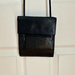 May include: A black leather crossbody bag with a flap closure and a zipped front pocket. The bag has a long, thin strap and is hanging against a white door. The bag appears to be new.