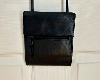 Könnte beinhalten: Eine schwarze Leder-Umhängetasche mit Klappenverschluss und einer Reißverschluss-Vordertasche. Die Tasche hat einen langen, dünnen Riemen und hängt an einer weißen Tür. Die Tasche scheint neu zu sein.
