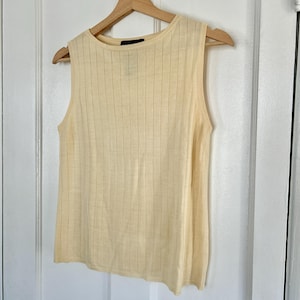 May include: A sleeveless, pale yellow ribbed knit top. The top is hanging on a wooden hanger against a white background. The fabric has vertical ribbing detail. The neckline is a simple, rounded scoop.