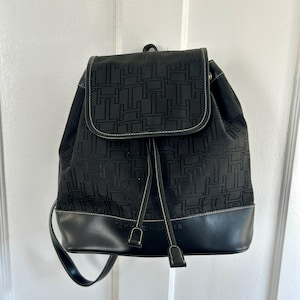 May include: Black backpack with a flap closure and drawstring. The backpack features a repeating pattern of the letter "H" and a black leather bottom. White stitching outlines the flap and the leather base. The backpack has adjustable straps.