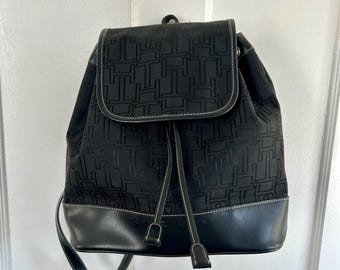 May include: Black backpack with a flap closure and drawstring. The backpack features a repeating pattern of the letter "H" and a black leather bottom. White stitching outlines the flap and the leather base. The backpack has adjustable straps.