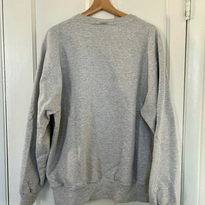 Pode incluir: Uma camisola cinza mesclada pendurada em um cabide de madeira contra uma porta branca. A camisola tem gola redonda, mangas compridas e punhos e bainha canelados. O tecido parece macio e confort&aacute;vel.