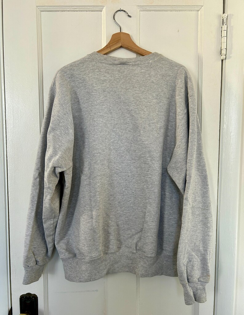 Pode incluir: Uma camisola cinza mesclada pendurada em um cabide de madeira contra uma porta branca. A camisola tem gola redonda, mangas compridas e punhos e bainha canelados. O tecido parece macio e confort&aacute;vel.