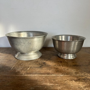 Könnte beinhalten: Zwei antike Metallschalen mit silbernem Finish. Die Schalen haben eine abgerundete Form und stehen auf einem Sockel. Die Schalen werden auf einer Holzoberfläche präsentiert.