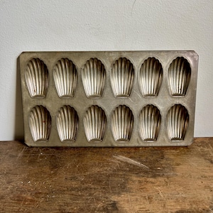 May include: A vintage metal baking pan with 12 individual moulds shaped like seashells. The pan is silver in colour and has a slightly worn appearance.