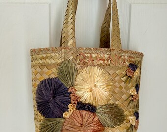 Könnte beinhalten: Eine kleine, gewebte Strohtasche mit einem braunen, beige und cremefarbenen Farbschema. Die Tasche hat ein gewebtes Design und verfügt über farbenfrohe, flauschige Verzierungen in Form von Blumen.