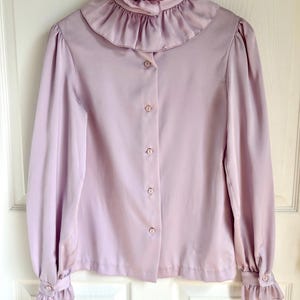 May include: A light purple blouse with a ruffled collar and cuffs. The long-sleeved shirt has a button-down front and is hanging on a wooden hanger. The fabric appears to be a soft, flowing material.